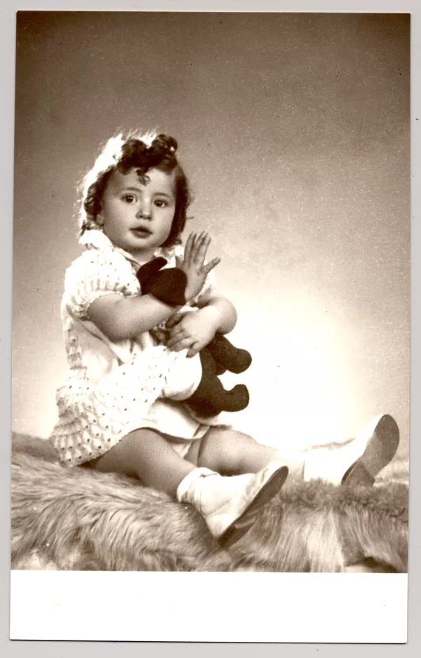 [Niñita de pelo rizado, vestido blanco abrazada a un juguete sentada sobre un felpudo]  [fotografía] Kyutaro Tunekawa.