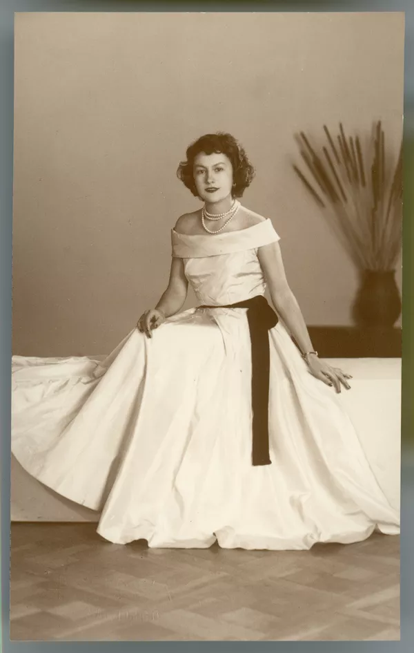 [Mujer posando con un vestido amplio y largo, hombros al desnudo, collar blanco de tres vueltas, sentada en una tarima y al fondo un macetero con espigas]  [fotografía] Kyutaro Tunekawa.