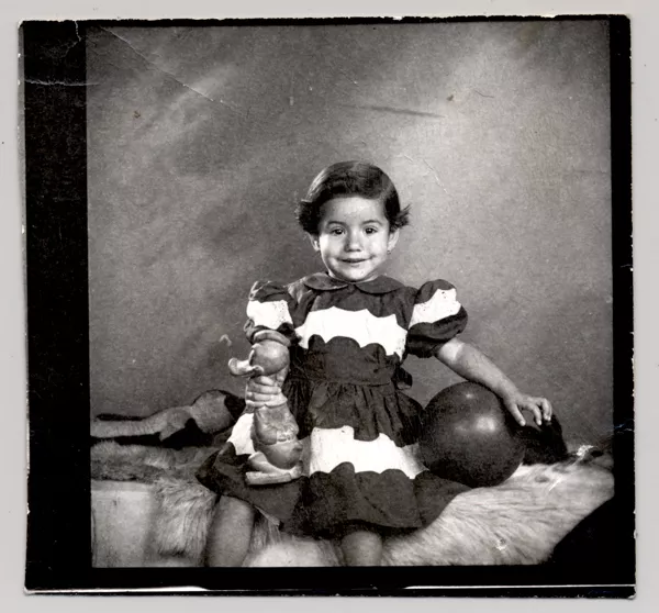 [Niñita de vestido rojo y blanco, con una pelota y un pato de juguete en sus manos mirando de frente]  [fotografía] Kyutaro Tunekawa.