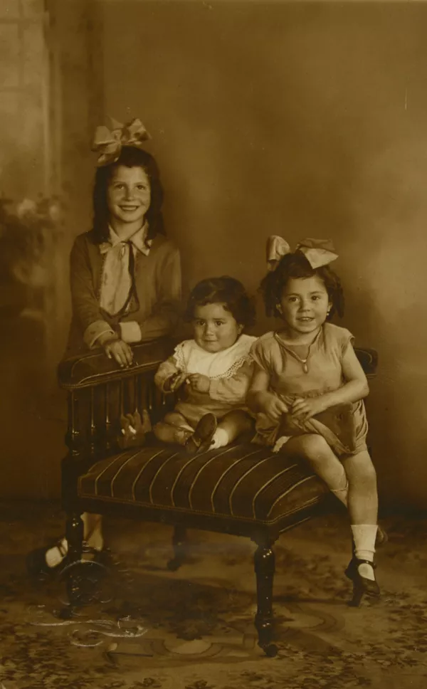 [Una niña de pie y las otras dos niñitas sentadas en un sillón]  [fotografía] Kyutaro Tunekawa.