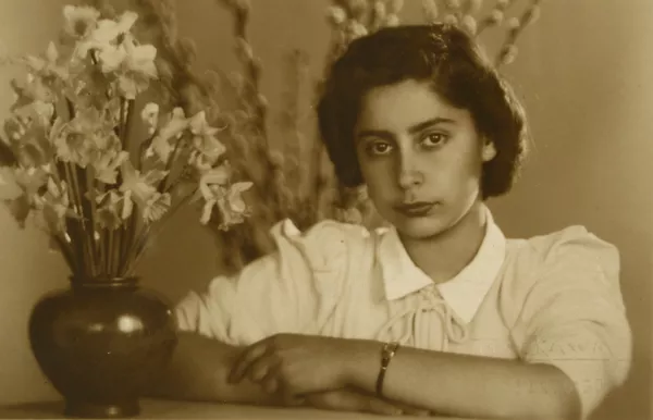 [Mujer joven, vestida de verano y apoyada en un mueble. En frente tiene un macetero con flores blancas y a sus espaldas se ve unos brotes de ramas largas]  [fotografía] Kyutaro Tunekawa.