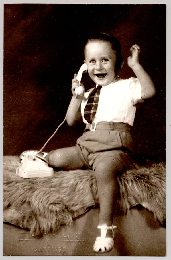 [Niño sonriente sentado sobre un felpudo con pantalones cortos, camisa blanca manga corta, corbatín a rayas y chalas blancas, simulando hablar por un teléfono de juguete]  [fotografía] Kyutaro Tunekawa.