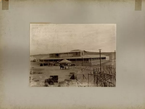 [Grupo de personas reunidas en el frontis de una oficina salitrera, vista de un poste con sombrilla para amarrar los caballos y un carruaje]  [fotografía].