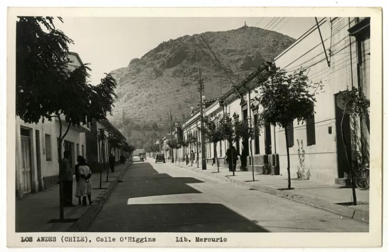 [Los Andes (Chile), Calle O'Higgins] : [Vista de la cumbre del Cerro Quicalcura, se divisa la imagen de la virgen; viviendas, vecinos conversando; en la esquina, un barrendero, autos de la época, un ciclista, tránsito mínimo] [fotografía] Col. Lib. Mercurio