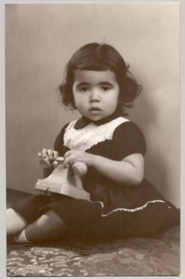 [Niñita morena de pelo corto con chasquilla sentada sobre una colchoneta forrada en brocato con un vestido oscuro y una cinta de broderí en el cuello y con un teléfono de juguete en sus manos]  [fotografía] Kyutaro Tunekawa.
